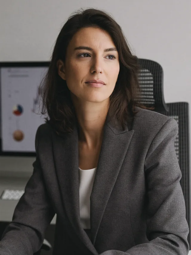 Finans uzmanı Elif arkasındaki ekranda grafik raporlarla masasında oturuyor